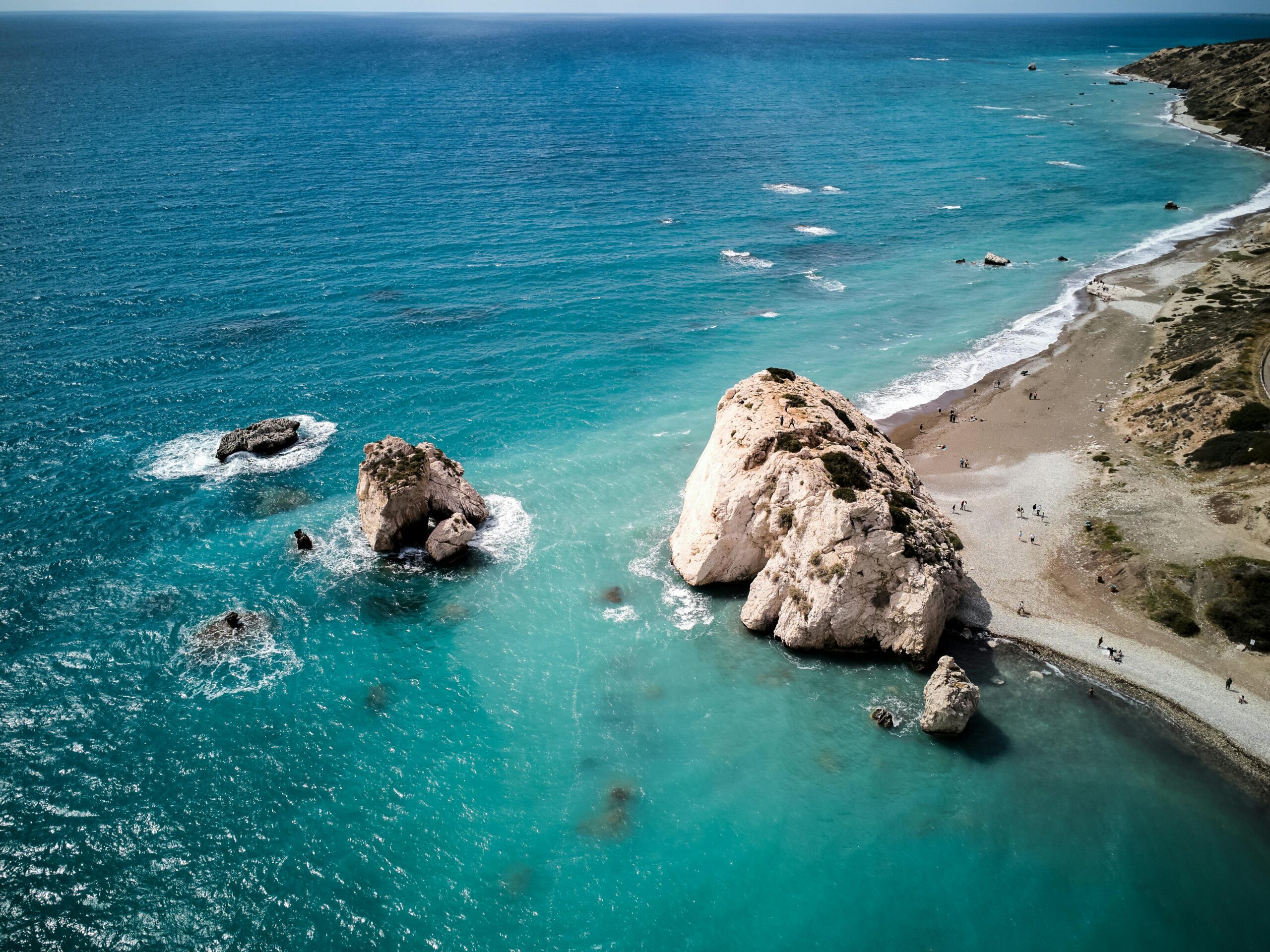 Stunning aerial shot of Petra tou Romiou rock formation along Cyprus coastline with azure sea and sandy beach.